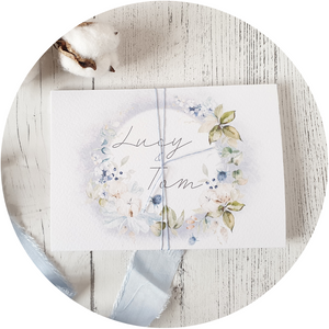 Dusty blue and floral wreath A6 Poppleberry all-in-one wedding invitation on white cardstock wrapped in dusty blue silk thread