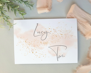 Close-up of a watercolour blush pink A6 Poppleberry accordion fold all-in-one wedding invitation, folded with gold sparkles