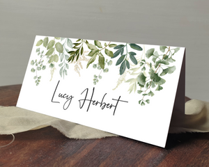 A Poppleberry greenery & eucalyptus wedding place card, printed with the guest's name, laid on a table & cloth.
