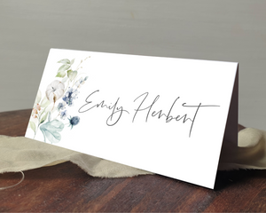 A Poppleberry dusty blue & cream cotton flowers wedding place card, printed with the guest's name, laid on a table & cloth.