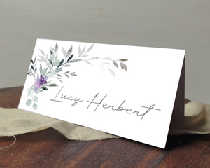 A Poppleberry lilac & grey flowers tent-fold wedding place card, printed with the guest's name, laid on a table & cloth.