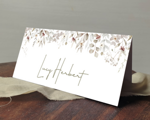 A Poppleberry neutral-shaded foliage wedding place card, printed with the guest's name, laid on a table & cloth.