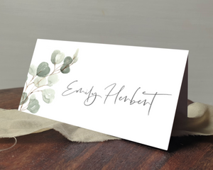 A Poppleberry muted green eucalyptus tent-fold wedding place card, printed with the guest's name, laid on a table & cloth.