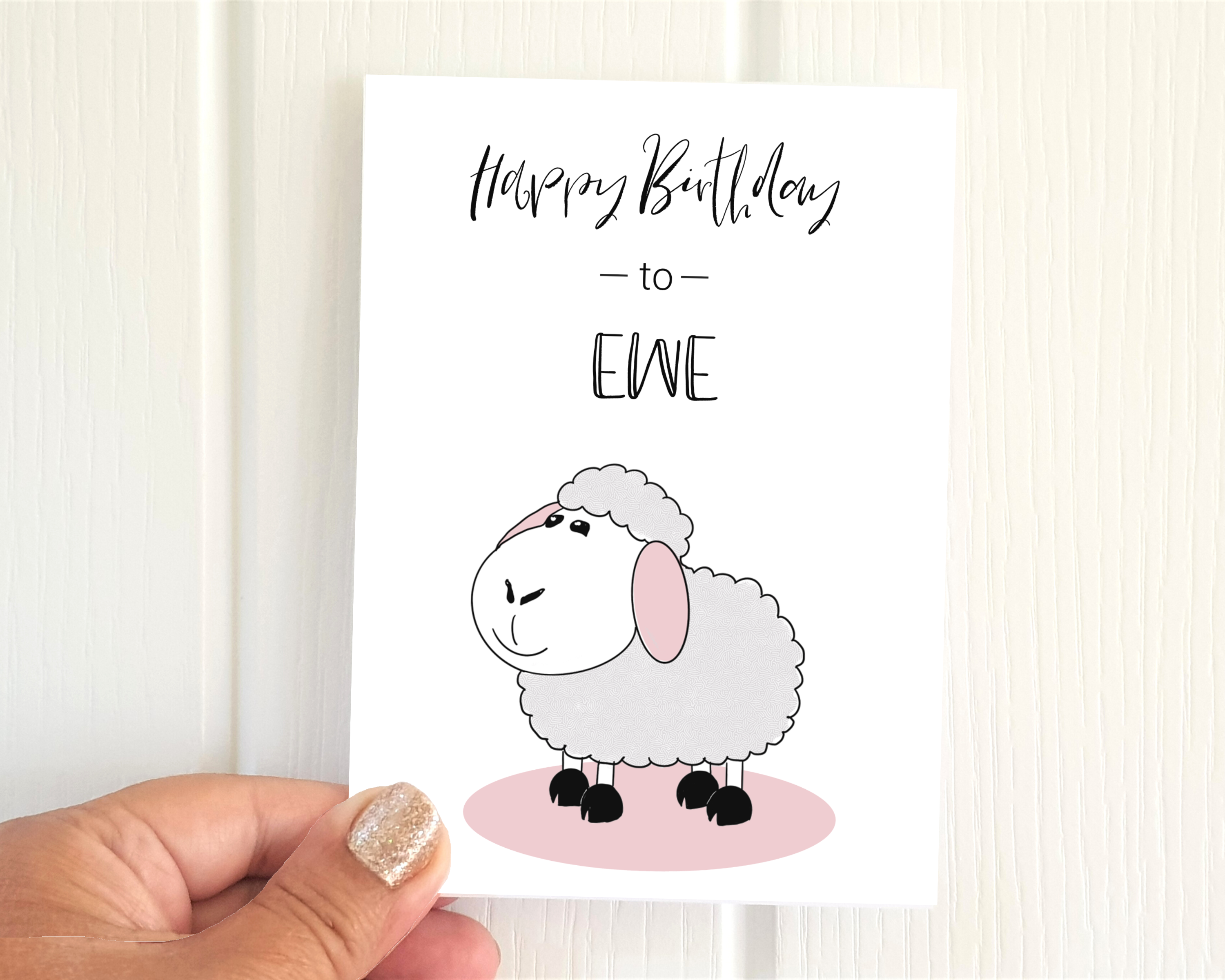 Poppleberry A6 folded birthday card, with a smiling white sheep illustration, on white cardstock, size compared to a thumb.