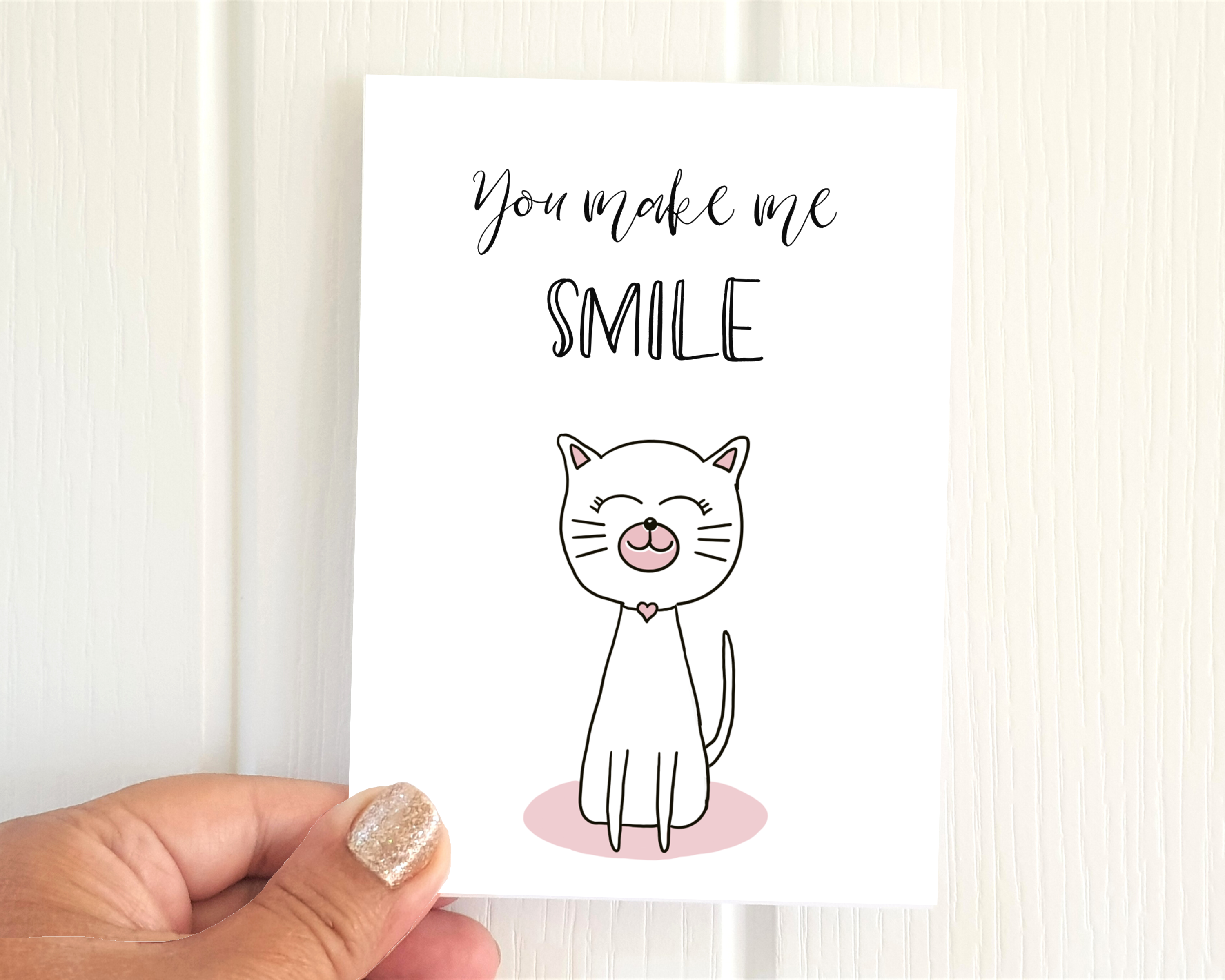 Poppleberry A6 folded greetings card, with a smiling white cat illustration, on white cardstock, size compared with hand.