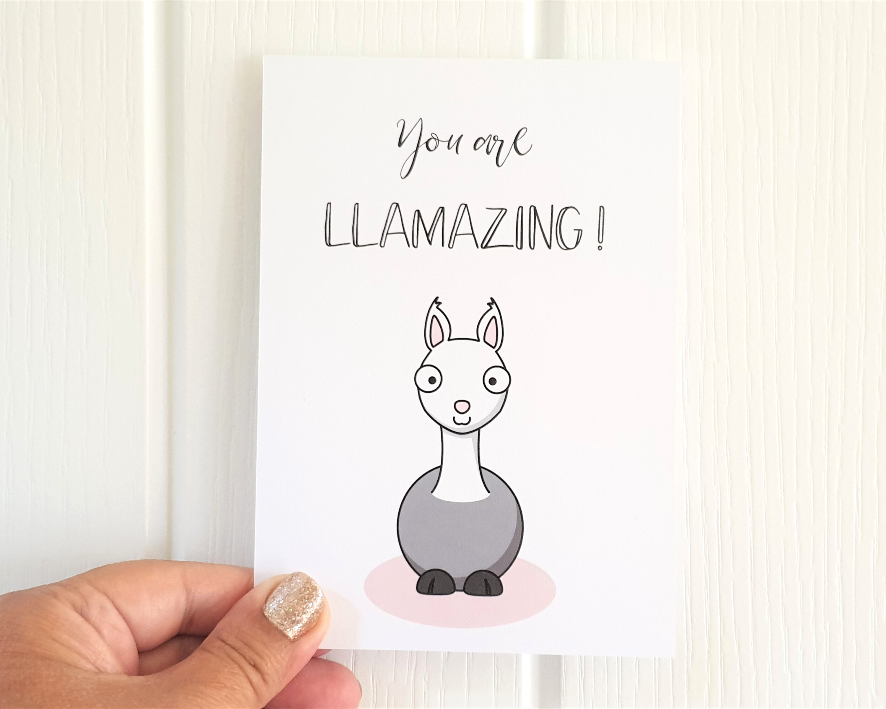 Poppleberry A6 folded greetings card, with a smiling grey llama illustration, on white cardstock, size compared with hand.